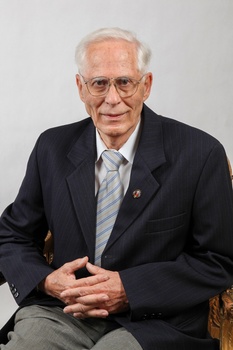 Idős férfi elegáns öltönyben ül egy széken Idős férfi elegáns öltönyben és nyakkendőben ül egy széken, kezei összekulcsolva.