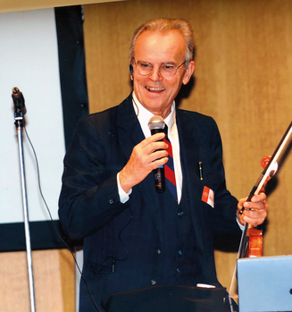 Elegáns férfi mikrofon és hegedű vonóval Elegánsan öltözött férfi mikrofont tart a kezében és hegedű vonót mutat, háttérben mikrofon állvány és vetítővászon.