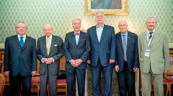 Elegáns férfiak csoportképe díszes teremben Hat elegánsan öltözött férfi áll egymás mellett egy díszes szobában, háttérben egy festmény.
