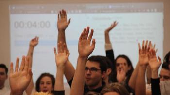 Csoport ember felemeli a kezüket egy előadás vagy szavazás során, háttérben kivetítőn időzítő és szöveg.