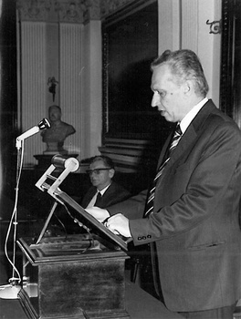 Férfi beszédet mond egy pulpitusnál elegáns teremben Férfi beszédet mond egy pulpitusnál, mikrofonnal, elegáns teremben, öltönyben.