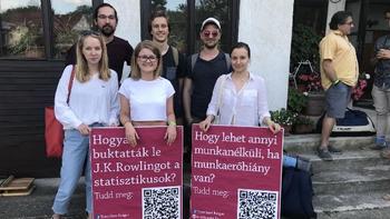 Fiatalok táblákat tartanak, melyeken kérdések és QR kódok szerepelnek, háttérben egy ház.