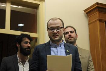 Hivatalos eseményen résztvevő emberek, egyikük papírt tartva beszédet olvas fel.