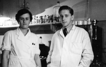 Laboratóriumi kutatás két személy részvételével Két személy laboratóriumi köpenyben egy laboratóriumban, üvegek és eszközök a háttérben.