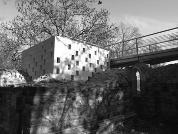 Modern geometrikus épület régi kőfal mellett, háttérben egy híd és fák láthatók.