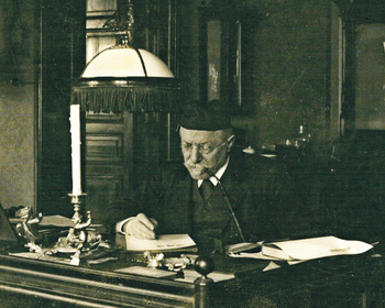 Régi íróasztal gyertyatartóval és asztali lámpával Régi íróasztal, rajta papírok, gyertyatartó és asztali lámpa egy klasszikus belső térben.