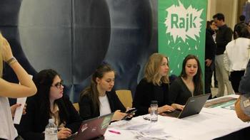 Rendezvény résztvevői Rajk bannerrel Emberek egy rendezvényen, asztalnál ülve, laptopokkal és papírokkal, háttérben Rajk banner.