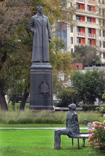 Szoborcsoport parkban modern épületekkel Szoborcsoport parkban, egy álló és egy ülő alak, háttérben modern épületek.