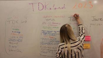Nő ír a fehér táblára, TDKaland 2023 felirat, nevek és témák láthatók.