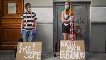 Két fiatal karton táblákkal tüntet, feliratokkal: 'free! SZFE' és 'KICSIT MÁR ELEGÜNK VAN'.